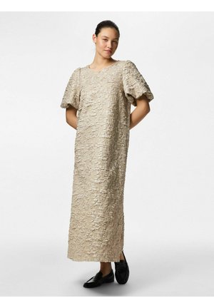 Vrouw staand met handen achter de rug, gekleed in een beige midi-jurk met structuur, pofmouwen en zwarte loafers op een effen witte achtergrond.