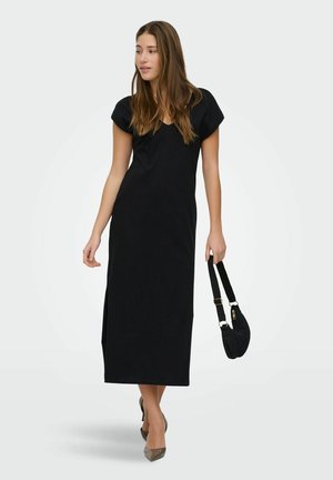 Ung kvinde iført en sort midi-kjole med korte ærmer, der holder en sort håndtaske og har brune spidse højhælede sko på, stående foran en ensfarvet baggrund.