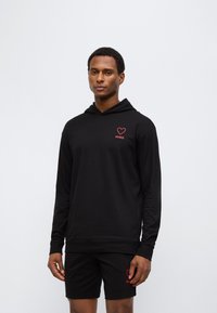 Μαύρο φούτερ με κουκούλα και μακριά μανίκια, με λογότυπο κόκκινης καρδιάς και κείμενο "HUGO". Συνδυάζεται με μαύρη σορτς που ταιριάζει, επίσης με το ίδιο λογότυπο.