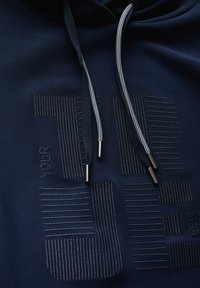 Mørkeblå hoodie med tekstureret design, prydet med ordene "DIN SELV" og stribede detaljer. Inkluderer matchende snore med metalspidser.