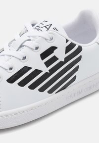Hvit sneaker med svarte diagonale striper, teksturert såle og stilig merking på siden. Har hvite snørebånd og glatt syntetisk materiale.