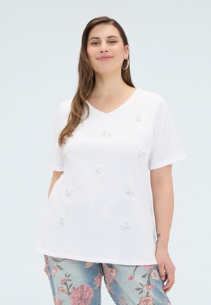 Kvinde iført en hvid T-shirt med V-hals med små blomsterbroderier og lyseblå jeans med store lyserøde blomsterprint, stående mod en lys baggrund.