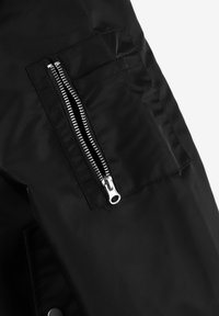 Czarna kurtka z wyraźnym srebrnym zamkiem błyskawicznym na kieszeni zapinanej na zamek, o gładkiej, matowej fakturze i minimalistycznym designie.