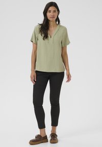 Blusa verde chiaro a maniche corte con scollo a V, texture a coste e cuciture decorative sul colletto; abbinata a pantaloni slim-fit neri.