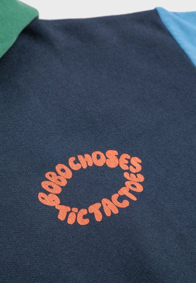 Marineblaues Polo mit grünem und hellblauem Kragen; verfügt über ein oranges, kreisförmiges Logo mit dem Text "Bobo Choses Tictactoe". Glatte Baumwollstruktur.
