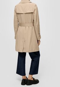 Osoba ubrana w beżowy trencz z paskiem, ciemne szerokie spodnie 3/4 oraz czarne mokasyny, stojąca na tle jasnym.