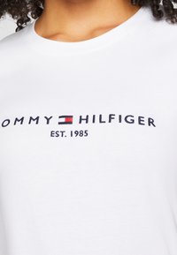 Biała koszulka z logo "Tommy Hilfiger" i haftowanym emblematem flagi powyżej napisu "EST. 1985" na piersi, noszona przez osobę z kręconymi włosami.