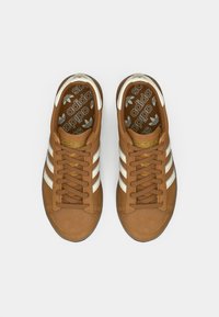 Brūni zamšādas sporta apavi ar baltām svītrām, noapaļota purngala forma un auklām. Adidas logo uz mēles un rakstainā iekšzole, plakana gumijas zole.