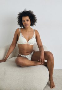 Soutien-gorge en dentelle crème et bikini assorti, avec des accents de dentelle florale, des bretelles réglables et une texture lisse. Modèle assis sur un canapé texturé.