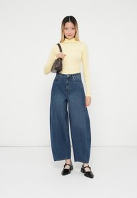 Top amarillo de manga larga, jeans de mezclilla azul de tiro alto y holgados, y zapatos de tacón negro clásicos. Bolso marrón de hombro con un asa en la parte superior.