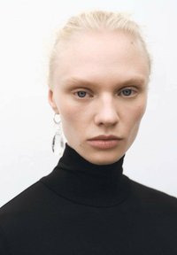 Eine Person mit einem schwarzen Rollkragenpullover, blasser Haut und blonden Haaren, trägt große, klare Tropfenohringe und hat einen neutralen Gesichtsausdruck.