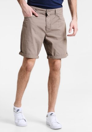 Mężczyzna ubrany w beżowe, podwinięte szorty, granatową koszulę oraz białe buty Nike, z jedną ręką w kieszeni na jednolitym tle.