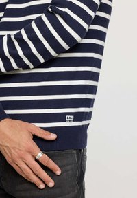 Pull-over en maille rayé bleu marine et blanc avec un ourlet et des poignets côtelés. Présente une étiquette de marque sur le bas à gauche. Modèle portant un jean noir.