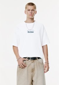 Ung mand iført en hvid oversized T-shirt med teksten "It's time", beige slidte jeans, sølv kæder og armbånd, stående foran en hvid baggrund.