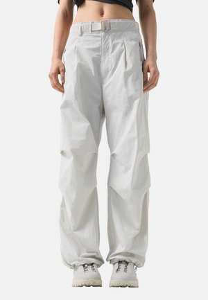 Osoba nosząca luźne białe spodnie cargo z paskiem i białe trampki, stojąca na jednolitym jasnym tle.