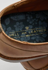 Interior de zapato de cuero marrón con un forro de patrón floral en azul oscuro, con texto dorado: "HELL FOR LEATHER NEXT" y una corona decorativa.