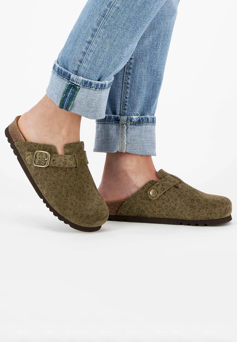 Olivgrüne Wildleder-Clogs mit Leopardenmuster, ausgestattet mit einer Metall-Schnalle und einer Korksohle. Die Hosenbeine sind hochgerollt, wodurch die Clogs sichtbar sind.