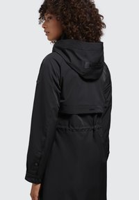 Giacca impermeabile nera con cappuccio, vita stretta e accenti di marca a contrasto sul retro. Il materiale è liscio con una finitura opaca.