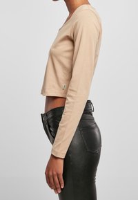 Beige Langarmshirt mit Rundhalsausschnitt, aus weichem Material, kombiniert mit hochgeschnittenen schwarzen Jacken aus Kunstleder mit Gesäßtaschen.