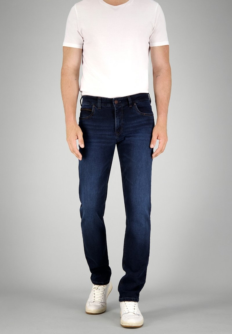 Donkerblauwe denim jeans met een slim fit, voorzien van een klassieke vijf-pocket ontwerp en contrasterende stiksels, gecombineerd met een wit T-shirt en sneakers.