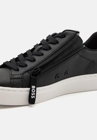Svart lädersneaker med dragkedja på sidan, platta svarta snörningar, texturerad vit gummisula och liten logotagg med ordet "BOSS."