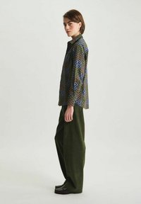 Chemise imprimée aux motifs verts, bleus et bruns ; pantalon en velours côtelé coupe décontractée de couleur vert foncé ; chaussures noires ; vue de profil.