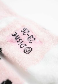 Calzini soffici rosa e bianchi con testo nero e motivo di faccine sorridenti; texture morbida con design accogliente; targhetta visibile in cima.