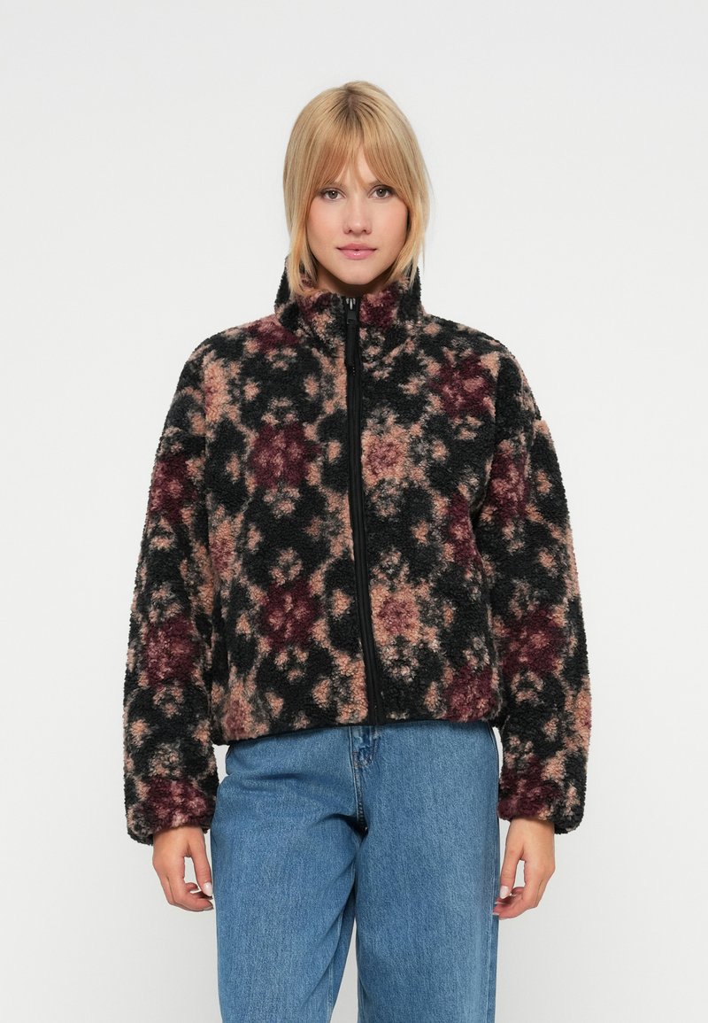 Chlupatá zipová bunda s černým, růžovým a bordó květinovým vzorem, vysokým límcem a volným střihem. Kombinována s světle modrými džíny.