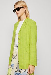 Limoengroene blazer van gestructureerde stof, met drie gouden knopen, een reverskraag en voorzakken, gecombineerd met een bloemenrok.