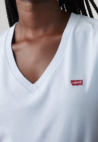 Světle modré bavlněné tričko s výstřihem do V a krátkými rukávy, s červenou nášivkou loga Levi's na levé oblasti hrudi. Hladká textura.