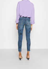 Pantalones vaqueros azules de denim con bordados florales en negro, de corte slim, con dobladillos deshilachados, combinados con una blusa lila, tiras en el tobillo y tacones de bloque.