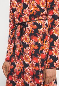 Gros plan d'une robe fleurie avec des fleurs rouges, orange et blanches sur un tissu noir, avec une fine ceinture noire nouée à la taille.