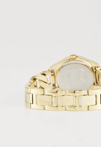 Montre en métal doré avec un boîtier rond élégant, un bracelet poli, et le logo gravé "GUESS" sur les maillons, mettant en valeur une surface brillante et réfléchissante.