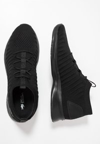 Chaussures de sport noires en maille avec une tige texturée, des lacets élastiques, et une semelle noire épaisse et coussinée avec un logo sur la semelle intérieure.