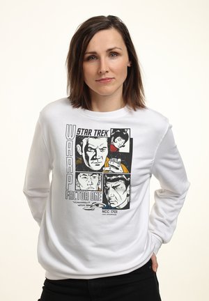 Kobieta o brązowych włosach do ramion nosi białą bluzę z czarno-białymi portretami w stylu komiksowym Star Trek oraz grafiką USS Enterprise.