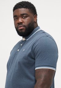 Man met een baard, gekleed in een blauwe polo met korte mouwen en een wit gestreepte kraag, met zijn gezicht iets schuin naar voren tegen een effen witte achtergrond.