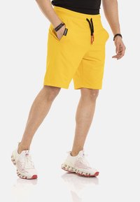 Gele shorts met een elastische tailleband en zwarte trekkoorden, met zijzakken. Draagt witte en rode sportschoenen.