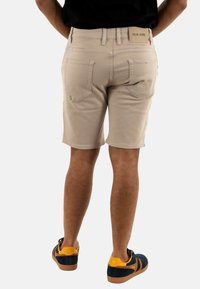 Pantaloni corti beige con una vestibilità slim, dotati di tasche posteriori cucite e un'etichetta in pelle sulla cintura. Abbinati a scarpe da ginnastica nere e arancioni.