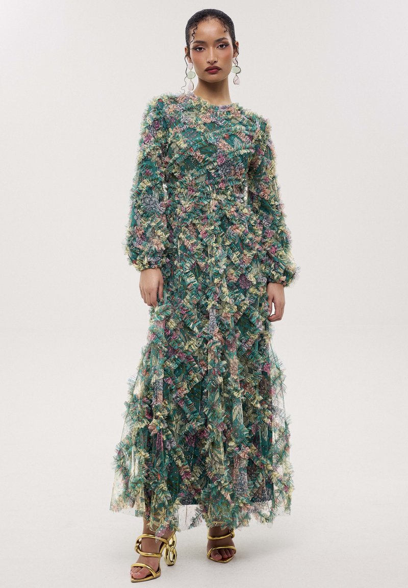 Woman standing, wearing a long-sleeve, full-length textured floral dress in green, pink, and blue tones with gold high-heeled sandals.