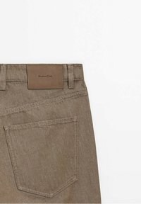 Section arrière d'un pantalon en denim marron montrant une poche unique et une étiquette de marque en cuir cousue sur la ceinture.