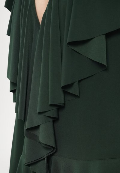 Ciemnozielona sukienka z warstwowymi falbanami, gładką fakturą i głębokim dekoltem w kształcie litery V. Tkanina elegancko opada na sylwetkę.