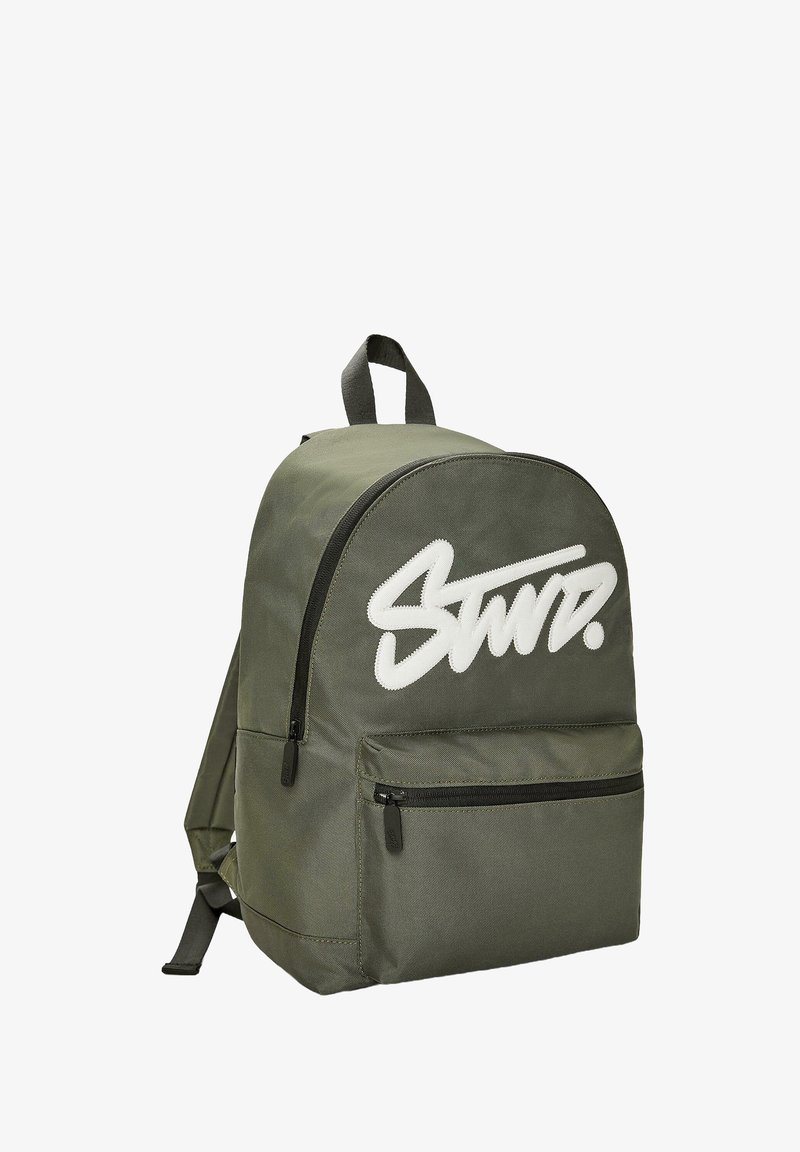 Olivazöld szövet hátizsák lekerekített tetejű, elülső cipzáras zsebbel és fehér hímzett logóval. Állítható pántokkal és akasztóval rendelkezik.