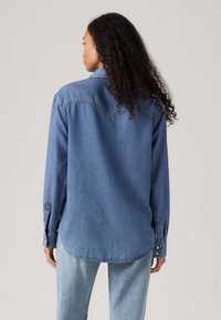 Dżinsowa koszula w jasnoniebieskim kolorze, z długimi rękawami, luźnym krojem, mankietami na guziki i zaokrąglonym dołem. Posiada tylną wstawkę oraz subtelne przeszycia.