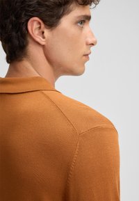 Polo in un tessuto a maglia color arancio bruciato. Presenta un colletto classico e maniche corte, con una texture sottile e una finitura liscia.