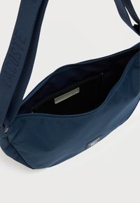 Tamnoplava crossbody torbica napravljena od izdržljive tkanine, s patentnim zatvaračem i unutarnjim džepom s mrežastim pregradnim dijelom. Prikazan je Lacosteov logo.