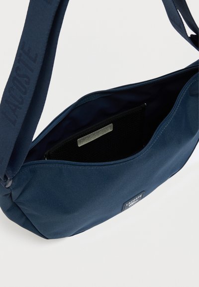 Sac bandoulière bleu marine en tissu durable, avec une fermeture éclair et une poche intérieure munie d'un séparateur en filet. Logo Lacoste visible.
