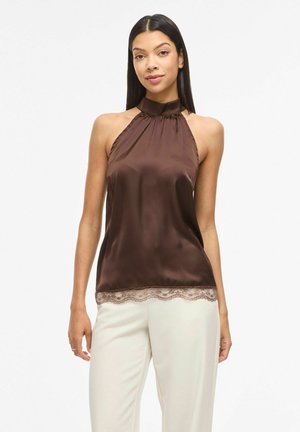 Kobieta w brązowym satynowym topie bez rękawów z koronkową lamówką, połączonym z białymi, wysokim stanem spodniami, stojąca na jednolitym tle.