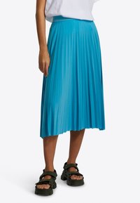 Mujer vestida con una falda midi plisada turquesa, una blusa blanca metida por dentro y sandalias negras de suela gruesa, de pie contra un fondo liso.