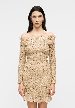 Kvinde med langt mørkt hår iført en beige, off-shoulder tekstureret kjole med flæsekant ved halsudskæring og skørt, stående mod en ensfarvet baggrund.