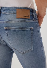 Skinny blue denim jeans with a light wash. Features a brown leather patch with embossed "mavi" logo at the waist and standard back pockets.
